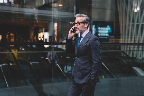 Elegante empresário de meia-idade com corte de cabelo na moda em óculos e traje preto falando no telefone em segundo plano urbano em Nova York - Foto, Imagem