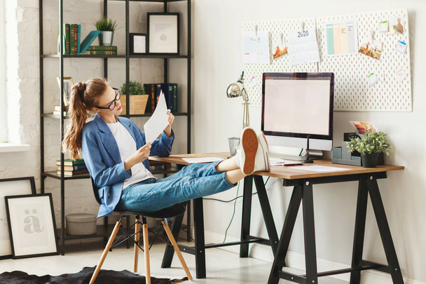 Оптимистичная молодая занятая женщина в повседневной одежде смотрит в камеру, сидя за столом с компьютером и бумагами и работая над проектом дома
 - Фото, изображение