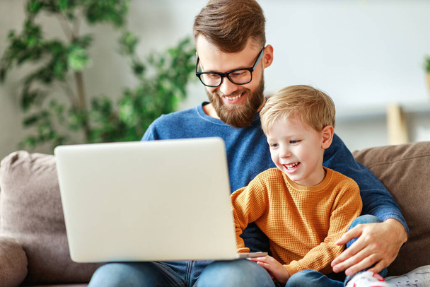 Cheerful bearded man in glasses hugging laughing boy and browsing laptop while sitting on sofa at home togethe - 写真・画像