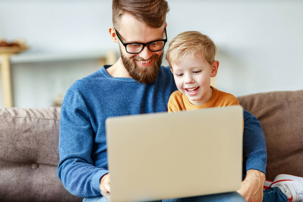 Cheerful bearded man in glasses hugging laughing boy and browsing laptop while sitting on sofa at home togethe - Foto, Bild