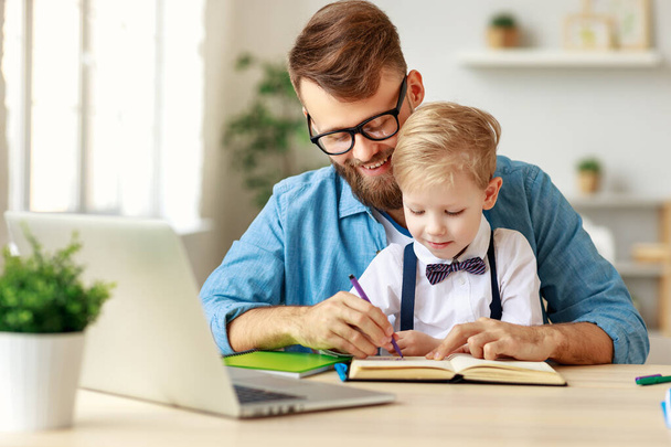 Молодой бородатый мужчина в очках помогает улыбающемуся маленькому мальчику с домашним заданием, проводя время вместе в доме
 - Фото, изображение