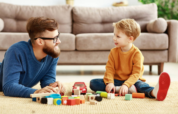 Вид сбоку симпатичного маленького мальчика и веселого молодого человека, проводящего свободное время вместе и играющего с разноцветными блоками на полу в гостиной roo
 - Фото, изображение