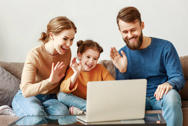 Positivos padres jóvenes amigables con una pequeña hija sonriente sentada en el sofá juntos respondiendo a una videollamada en el ordenador portátil y saludando con la mano mientras se relaja en casa en weeken
 - Foto, Imagen
