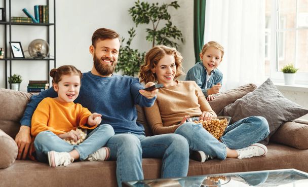 Позитивные улыбающиеся родители сидят на удобном диване с маленьким сыном и дочерью, наслаждаясь фильмом и поедая попкорн в доме
 - Фото, изображение