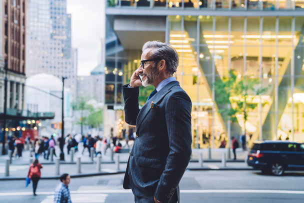 Side view of mature male in dark business suit talking on phone with smile in New York street and putting hand in pocket - Zdjęcie, obraz