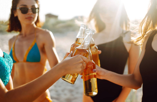 Chicas delgadas aplauden y beben cervezas en la playa al atardecer. Joven chica de cuatro en bikini disfrutando de unas vacaciones en la playa. Vacaciones de verano, vacaciones, relax y estilo de vida
. - Foto, imagen