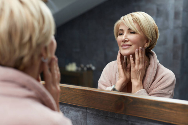 Skincare Routine. Mature Woman Touches Face Skin In Front Of The Mirror Reflection. Anti Aging Beauty Treatment Concept. - Foto, imagen