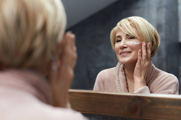 Skincare Routine. Mature Woman Uses Cosmetic Cream On Face Skin In Front Of The Mirror Reflection. Anti Aging Beauty Treatment Concept. - Photo, Image