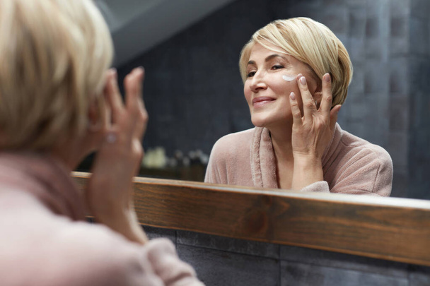 Skincare Routine. Mature Woman Uses Cosmetic Cream On Face Skin In Front Of The Mirror Reflection. Anti Aging Beauty Treatment Concept. - Foto, imagen