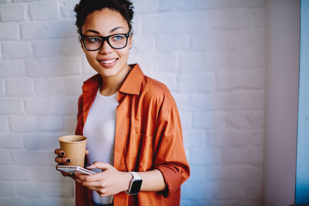 Positive female blogger holding paper cup with caffeine beverage and waiting friend indoors while chatting with followers on smartphone gadget, happy hipster girl in eyewear standing with cellular - Foto, Imagem