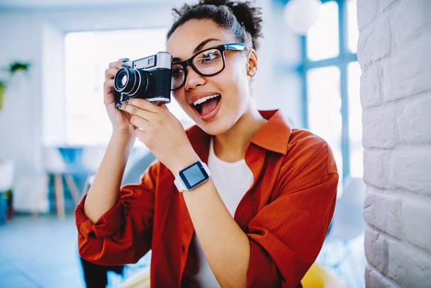 Surprised female amateur photographer in optical spectacles making photo via vintage camera, dark skinned amazed woman with expression on face taking pictures using old fashioned equipment indoors - Fotoğraf, Görsel