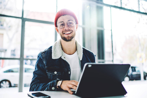 Cheerful young stylish male freelancer in warm clothes and glasses sitting at table with gadgets smiling at camera on blurred background - Φωτογραφία, εικόνα