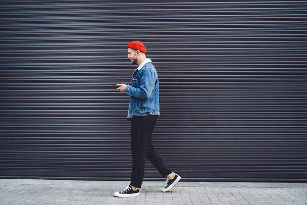 Vista lateral de un hombre joven y elegante con gorra roja y chaqueta de mezclilla caminando por la calle y utilizando el teléfono móvil como GPS contra el fondo del garaje
 - Foto, Imagen