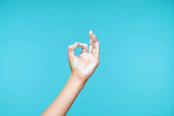 Photo of painted in white color hand being raised while showing mudra gesture, posing over blue background while meditating. Body language concept - Photo, Image