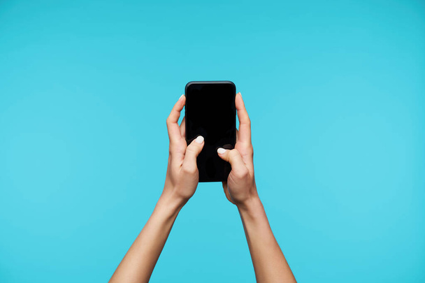 Indoor shot van mooie dame de handen houden van mobiele telefoon en houden duimen op zwart scherm, het typen van tekst op virtueel toetsenbord, terwijl geïsoleerd over blauwe achtergrond - Foto, afbeelding