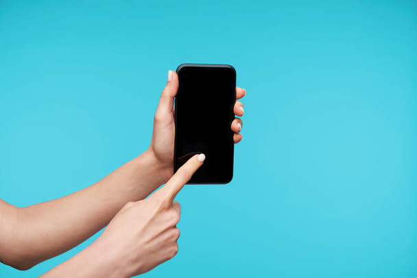 Κοντινό πλάνο του μαύρου σύγχρονου smartphone που διατηρείται από τα χέρια της γυναίκας με λευκό μανικιούρ, συγκινητική οθόνη με δείκτη, ενώ στέκεται πάνω από το μπλε φόντο - Φωτογραφία, εικόνα