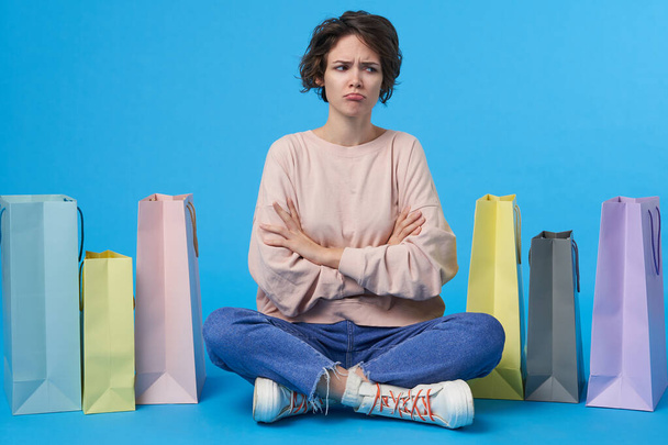 Offended young brown-eyed curly brunette woman frowning eyebrows and crossing hands on chest while sitting over blue background with multi-colored paper bags - Photo, Image