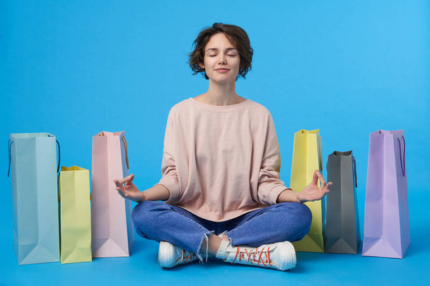 Pleasant looking young cute short haired brunette female meditating with closed eyes and forming fingers in mudra gesture, sitting over blue background with purchases - Photo, Image