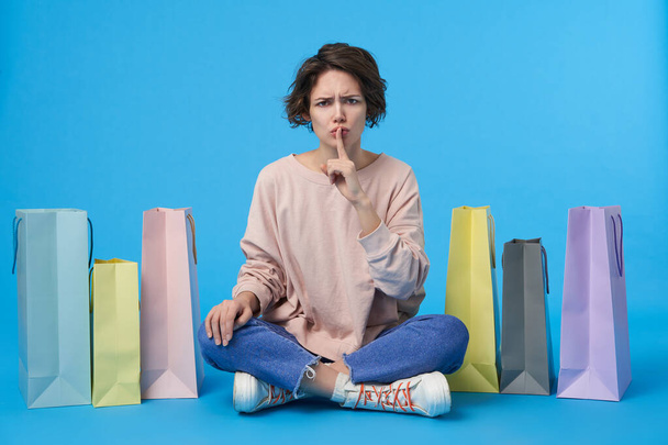 Serious young pretty dark haired curly lady with natural makeup keeping raised forefinger on her mouth while looking severely at camera, sitting over blue background with crossed legs - Photo, Image