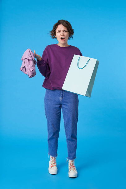 Disgruntled young lovely dark haired curly lady with natural makeup looking at camera with pout while keeping clothes and paper bag in raised hands, isolated over blue background - 写真・画像