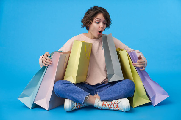 Amazed young curly brunette woman with short haircut holding raised hands on paper bags and looking excitedly at it, sitting over blue background in casual wear - Valokuva, kuva