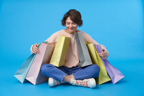 Good looking young dark haired curly lady with casual hairstyle smiling pleasantly while sitting over blue background with purchases, being in nice mood after shopping day - Photo, Image