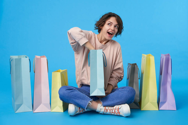 Cheerful young short haired curly female with casual hairstyle looking wonderingly what is inside of paper bag in her hands, isolated over blue background - Photo, Image