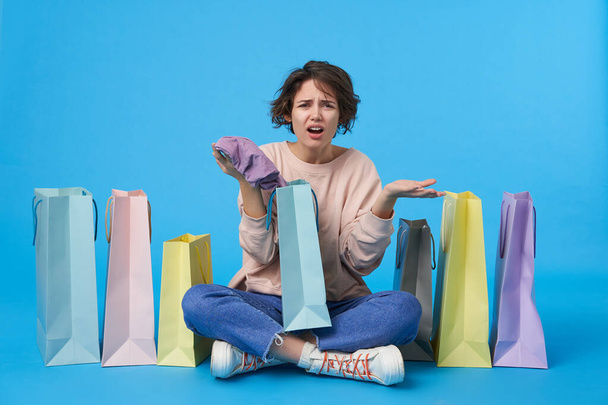 Displeased young pretty dark haired curly lady with casual hairstyle raising discontentedly palms and frowning her face while sitting over blue background with paper bags - Photo, Image
