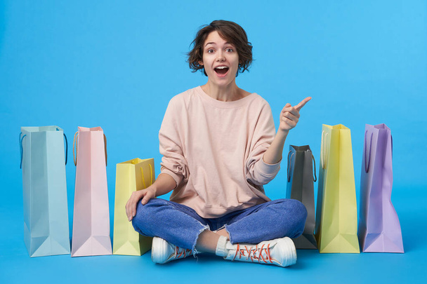 Overjoyed young lovely dark haired curly lady with casual hairstyle looking excitedly at camera with wide smile and pointing upwards while sitting over blue background with paper bags - Photo, Image