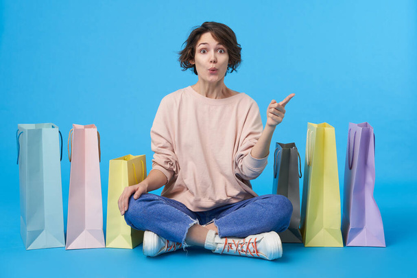 Excited young short haired female with natural makeup rounding surprisedly her brown eyes while pointing upwards with forefinger, sitting over blue background with crossed legs - Photo, Image