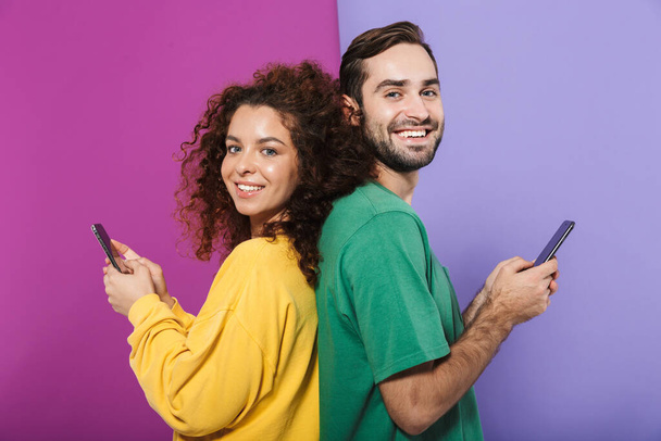 Portrait of positive caucasian couple in colorful clothing smiling and holding cellphones standing back to back isolated over violet background - Фото, изображение