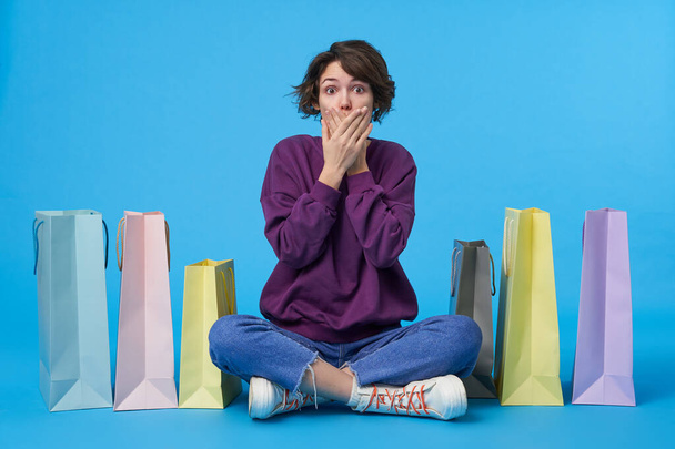 Shocked young brown-eyed short haired curly lady covering her mouth with hands and raising surprisedly eyebrows while looking at camera, isolated over blue background - Photo, Image