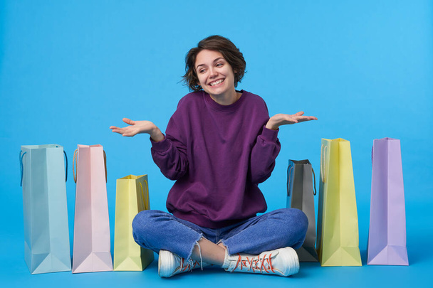 Positive young lovely dark haired woman with casual hairstyle shrugging with raised palms and smiling cheerfully, dressed in purple sweatshirt, jeans and sneakers while sitting over blue background - Photo, Image