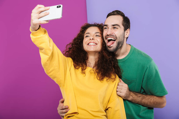 Portrait of positive caucasian couple in colorful clothing having fun while taking selfie photo on cellphone isolated over violet background - 写真・画像