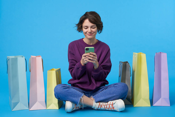 Pozytywny młody atrakcyjny kręcone brunetka kobieta trzyma telefon komórkowy w podniesionych rękach i patrząc radośnie na ekranie, siedzi na niebieskim tle z papierowymi torbami - Zdjęcie, obraz