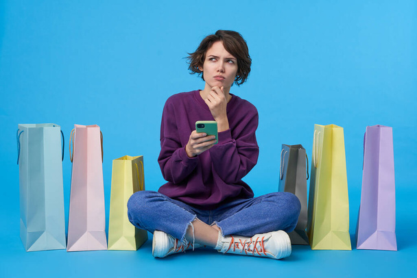 Προσεκτική νεαρή καστανομάτα σγουρά μελαχρινή γυναίκα που κρατάει smartphone και κατσουφιασμένα φρύδια ενώ κοιτάζει προς τα πάνω, απομονωμένη σε μπλε φόντο με αγορές - Φωτογραφία, εικόνα