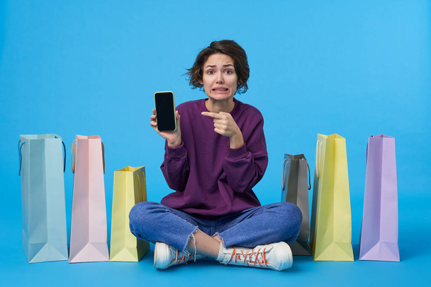 Bemused jonge vrij korte haren krullende vrouw houdt mobiele telefoon in opgeheven hand en toont verwarrend op het scherm, poseren over een blauwe achtergrond met gekruiste benen - Foto, afbeelding