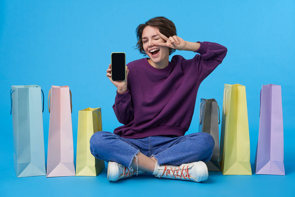 Cheerful young pretty dark haired curly female showing screen of her phone to camera and raising hand with victory gesture, sitting over blue background with paper bags - Photo, Image