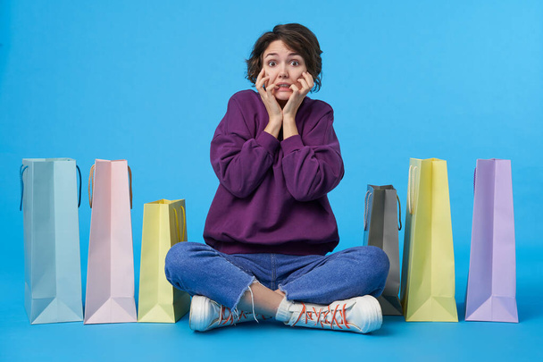 Frightened young dark haired curly female with short haircut holding her face with raised hands and looking scaredly at camera, sitting over blue background with crossed legs - Фото, изображение
