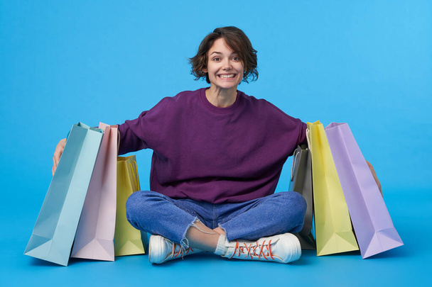 Indoor shot of glad pretty young brunette female with short haircut embrcing bags with her purchases and smiling widely at camera, isolated over blue background - Photo, Image