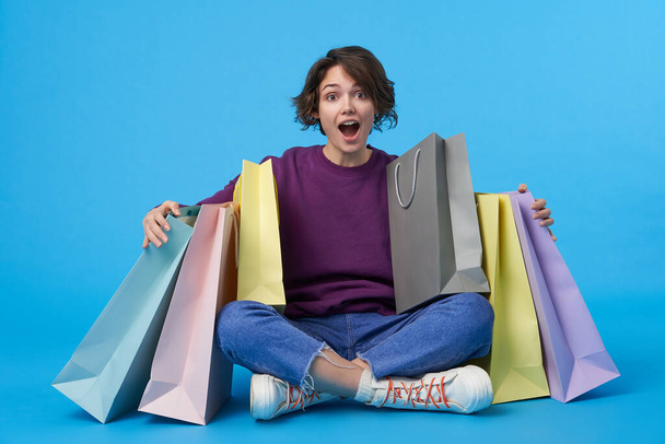 Overjoyed young attractive curly brunette woman embracing her purchases and keeping her mouth wide opened while looking heatedly at camera, posing over blue background - Foto, Imagen