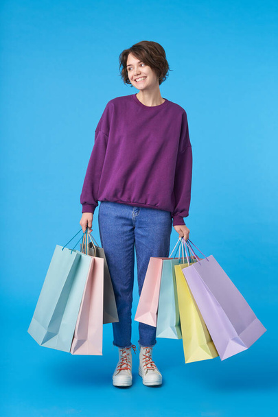 Indoor photo of good looking young glad dark haired curly woman dressed in casual clothes smiling pleasantly while standing over blue background with her purchases - Foto, Imagem