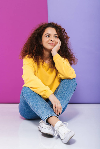 Full length image of attractive happy curly woman swearing casual clothes smiling while sitting on floor isolated over purple background - Foto, immagini
