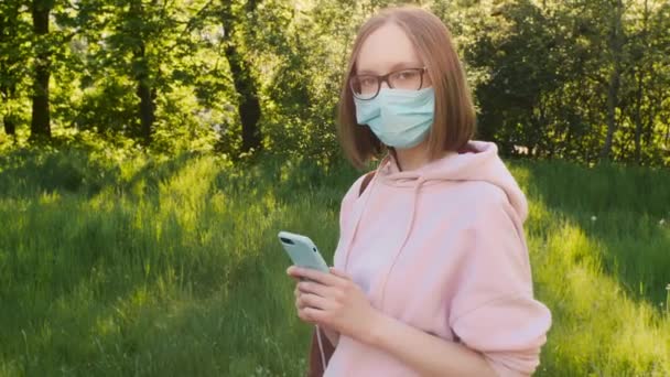 Portret van een jong trendy meisje in een medisch beschermend masker met een smartphone in het park. - Video