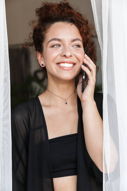 Image of a redhead positive smiling young lady in lingerie swimwear posing indoors in home hotel near curtain talking by mobile phone. - Fotografie, Obrázek