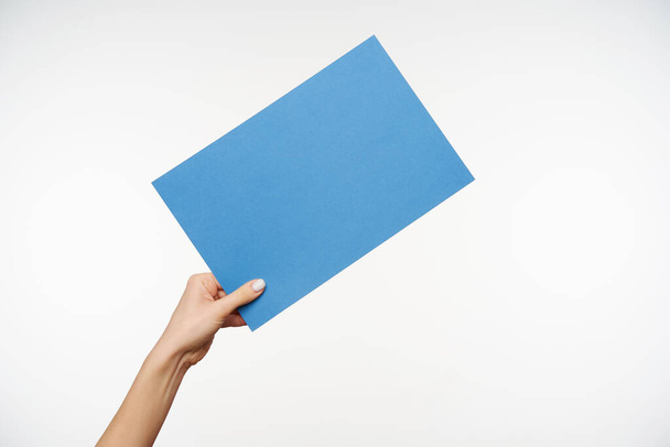 Indoor shot of young female's elegant hand being raised while keeping piece of light blue paper in it, passing it to someone while posing over white background - Photo, Image