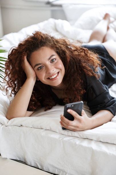 Εικόνα μιας κοκκινομάλλας αισιόδοξης νεαρής με εσώρουχα σε εσωτερικούς χώρους στο σπίτι ξενοδοχείο στο κρεβάτι χρησιμοποιώντας το κινητό τηλέφωνο. - Φωτογραφία, εικόνα