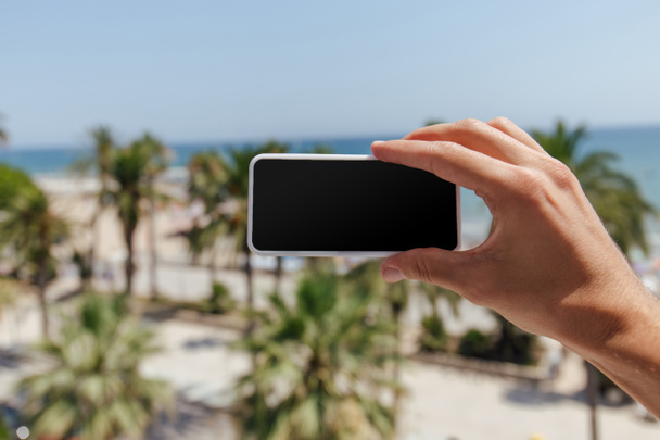 Cropped view of man holding smartphone with blank screen with sea coast at background - Photo, Image