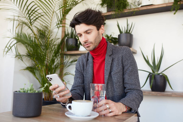 Sérieux jeune homme d'affaires attrayant aux cheveux bruns vêtu de vêtements élégants vérifiant sa boîte aux lettres sur smartphone en attendant quelqu'un dans un café de la ville
 - Photo, image
