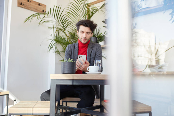 Indoor shot of young pretty dark haired bearded guy with trendy haircut sitting over modern cafe interior and looking on his smartphone, waiting for someone while having meeting out of office - Foto, Imagem